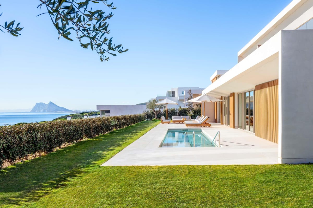 Un área de piscina exterior moderna con sillones de descanso, vista despejada al mar y un césped bien cuidado.