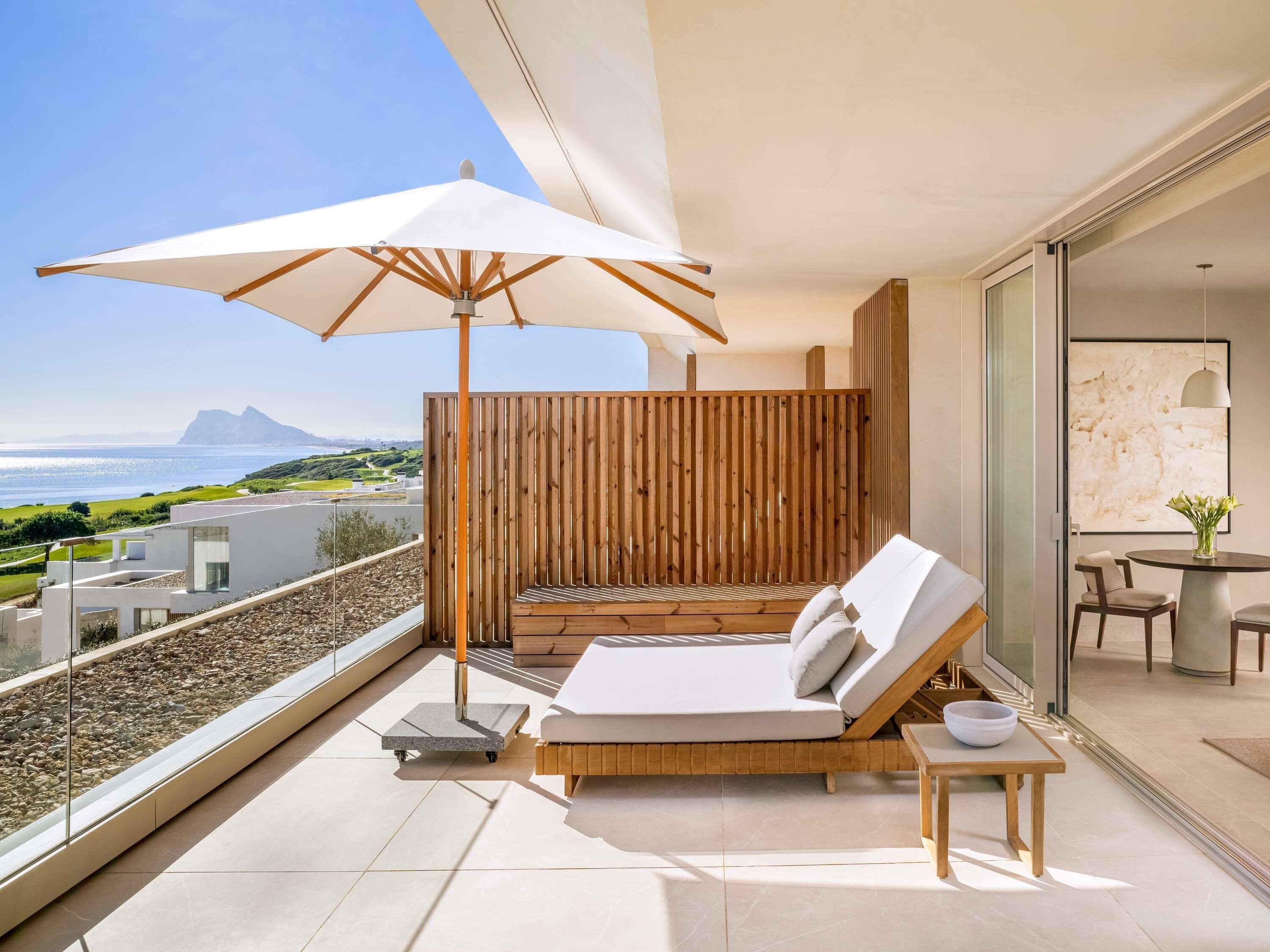 A balcony with a white umbrella, a lounge chair, a small table, and a view of the sea and landscape.