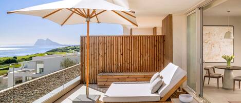 A balcony with a white umbrella, a lounge chair, a small table, and a view of the sea and landscape.