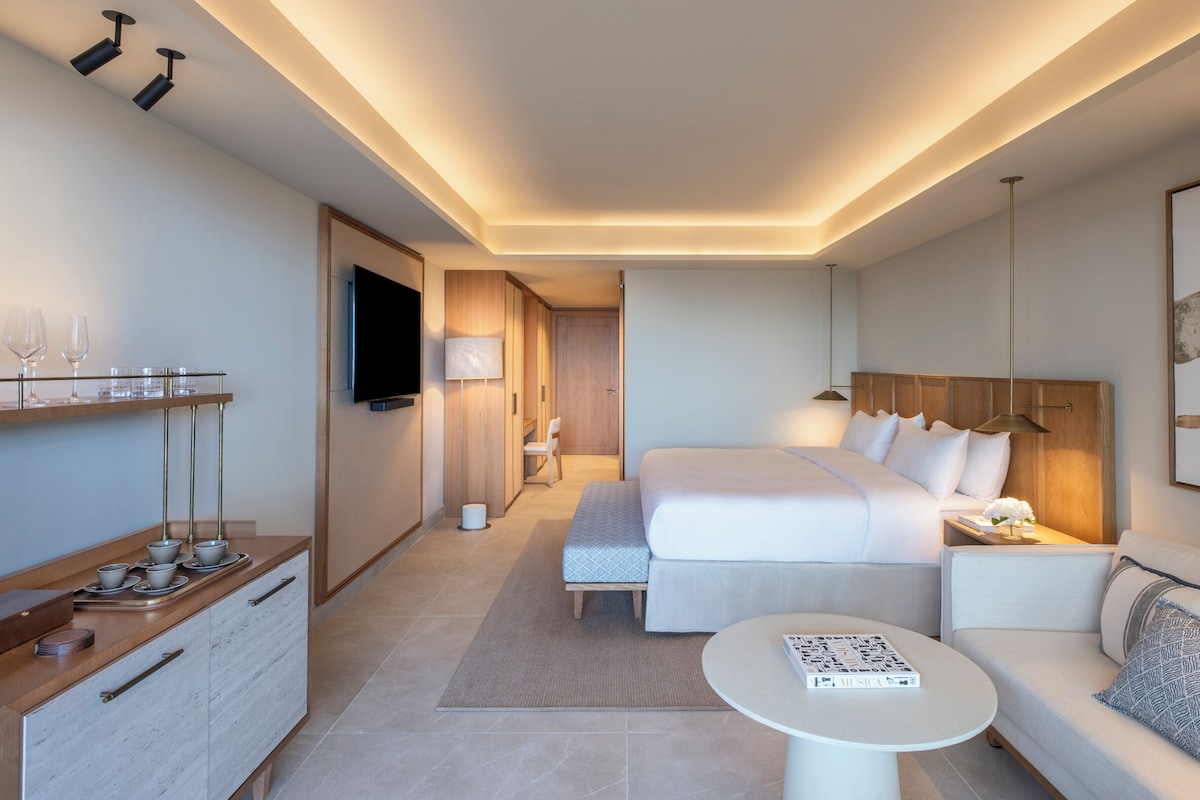 A modern hotel room with a large bed, a sofa, a small table, a TV, and a wooden cabinet with cups and glasses.