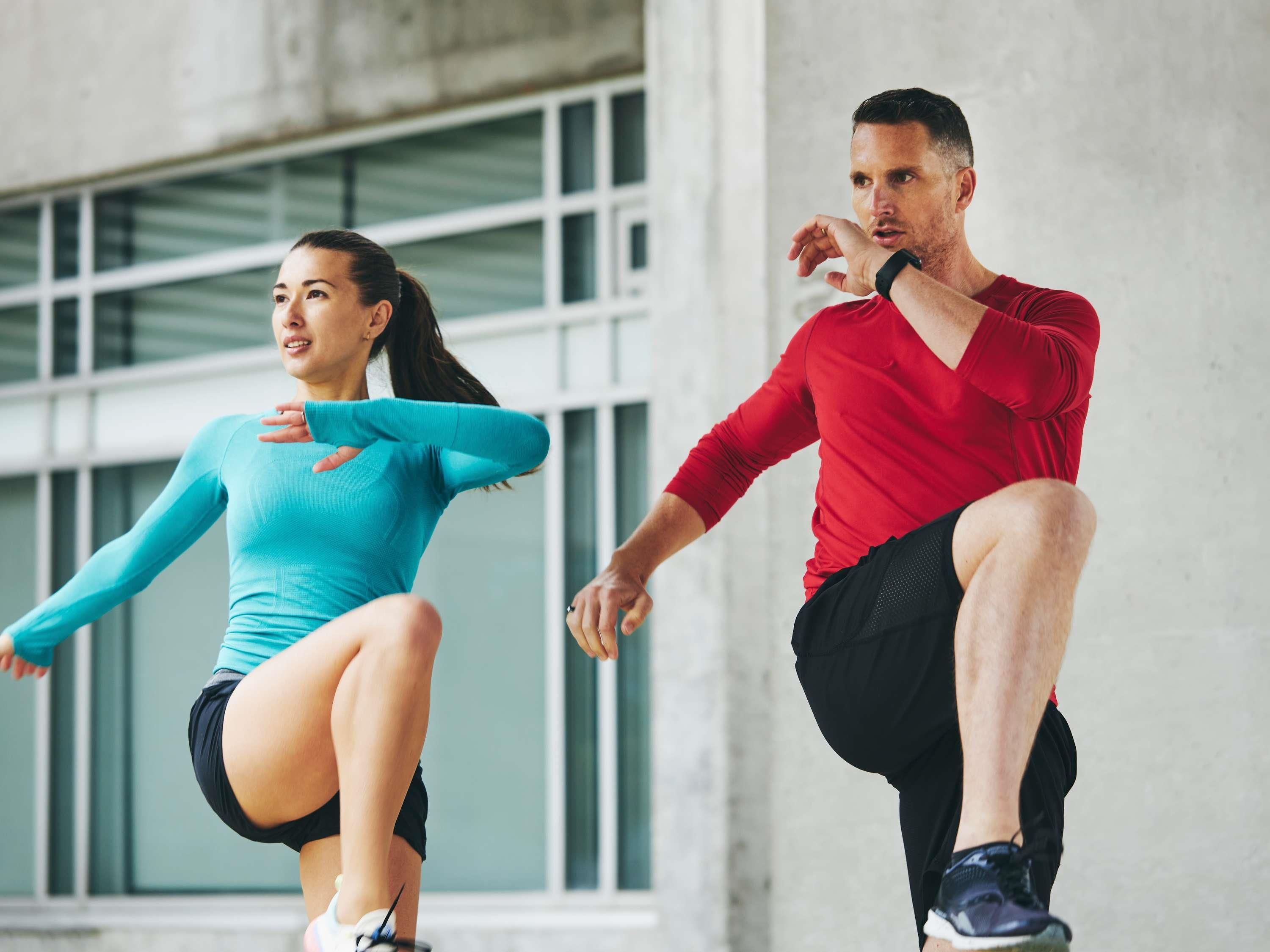 Dos personas con ropa deportiva, una con camiseta verde azulado y pantalones cortos negros, y la otra con camiseta roja y pantalones cortos negros, realizando un ejercicio de sentadilla.