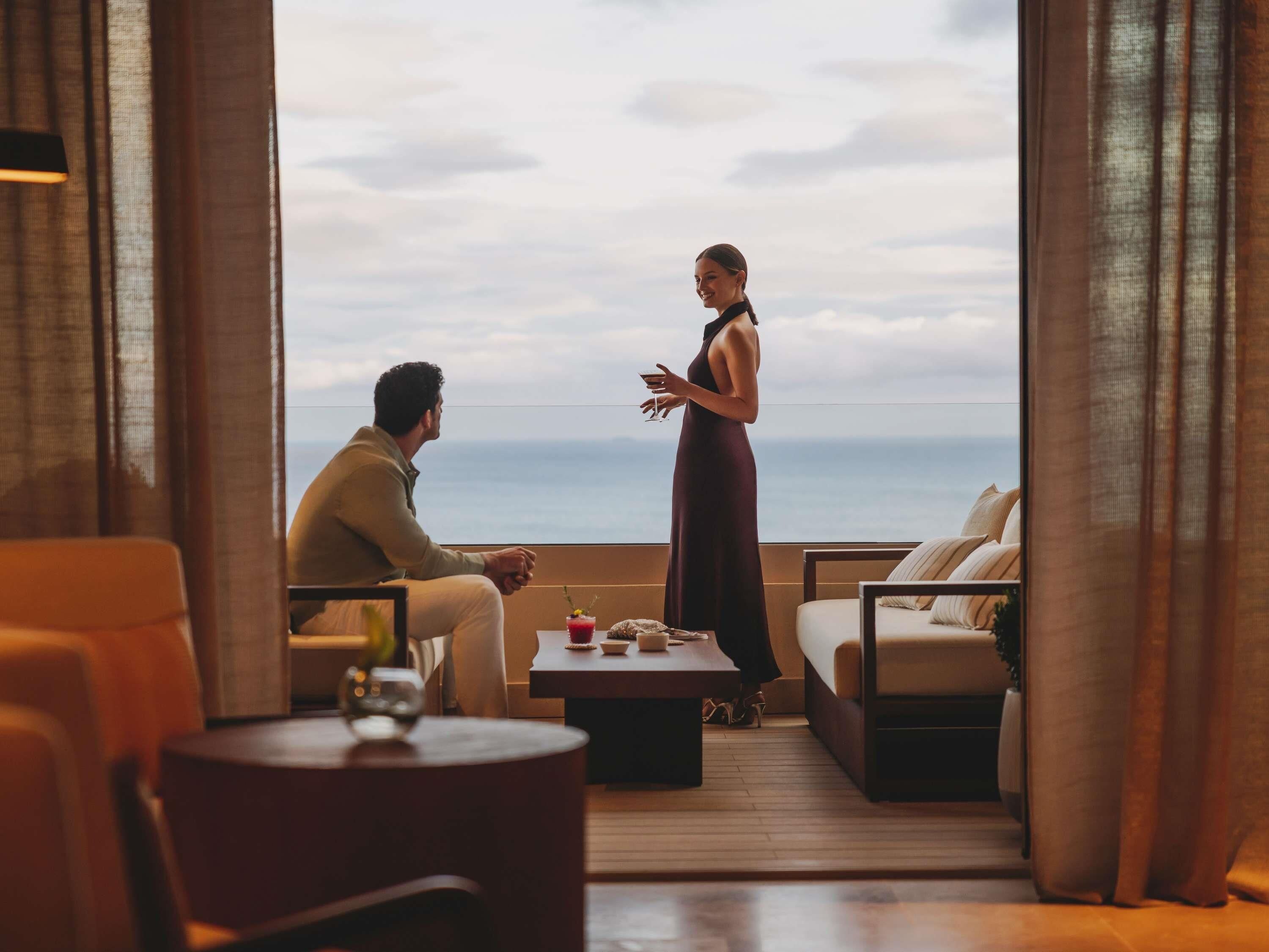 Une pièce avec vue sur la mer, un canapé, une table avec des boissons et une personne debout.