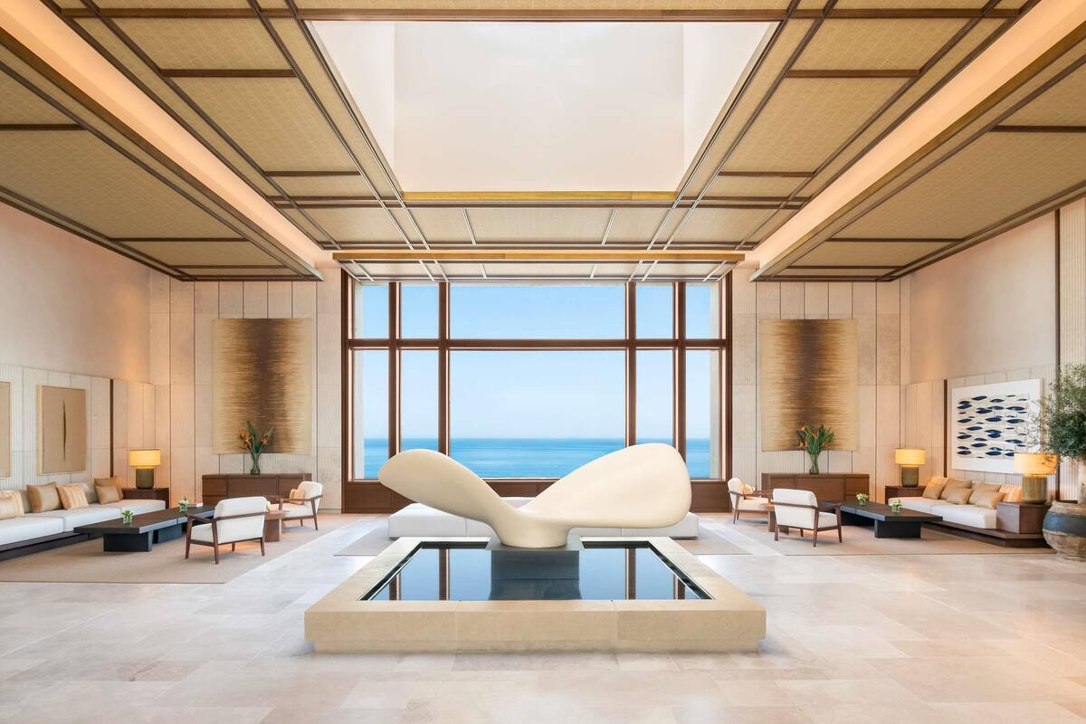 A spacious lobby with a large window overlooking the sea, a central seating area, and a modern design with wooden paneling and a high ceiling.