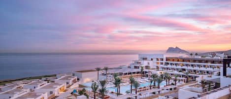 A resort with multiple buildings, a pool area, and a view of the sea at sunset.