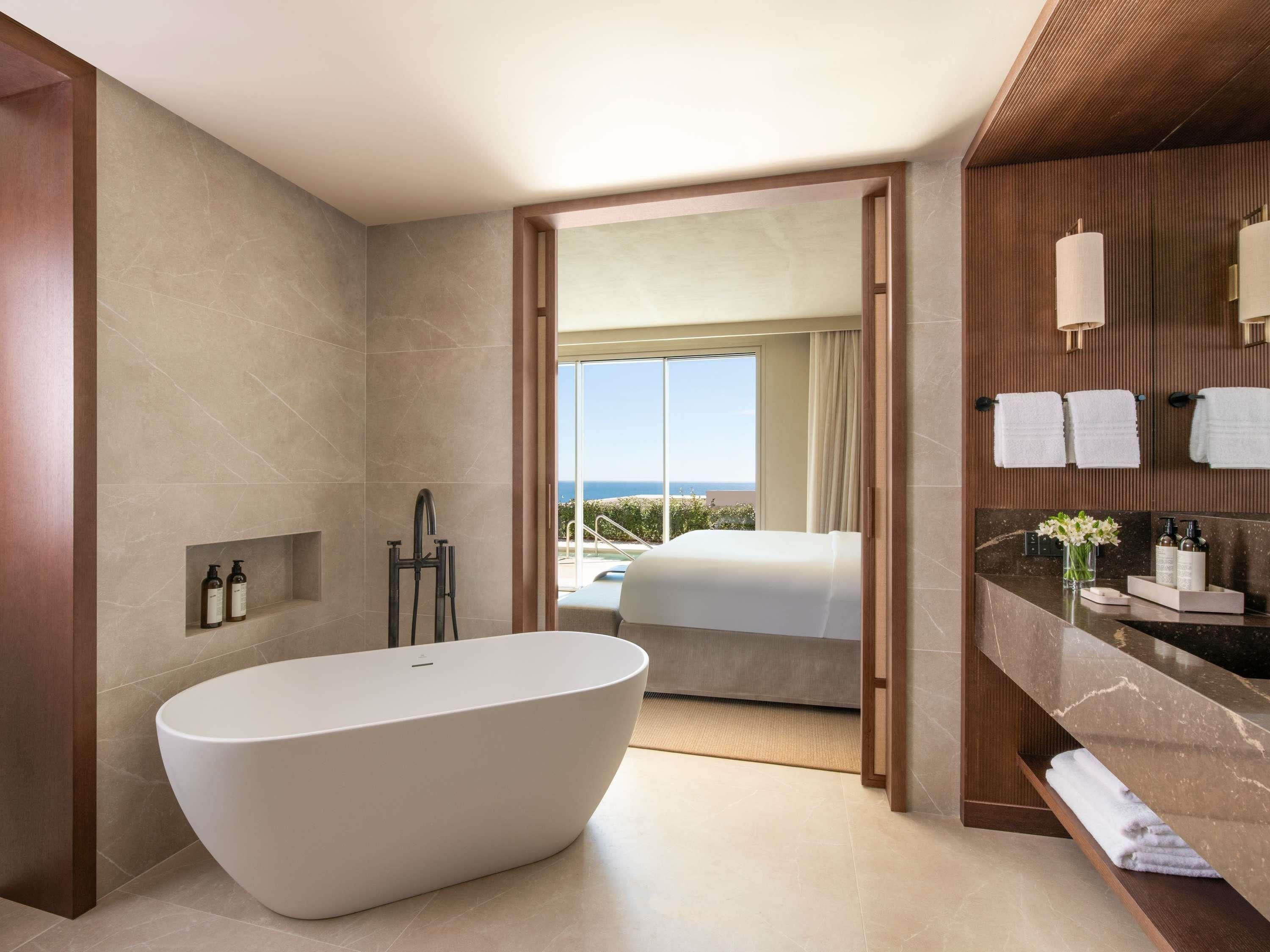 Un baño moderno con una bañera independiente, un tocador con encimera de mármol y vistas a un balcón con cama.