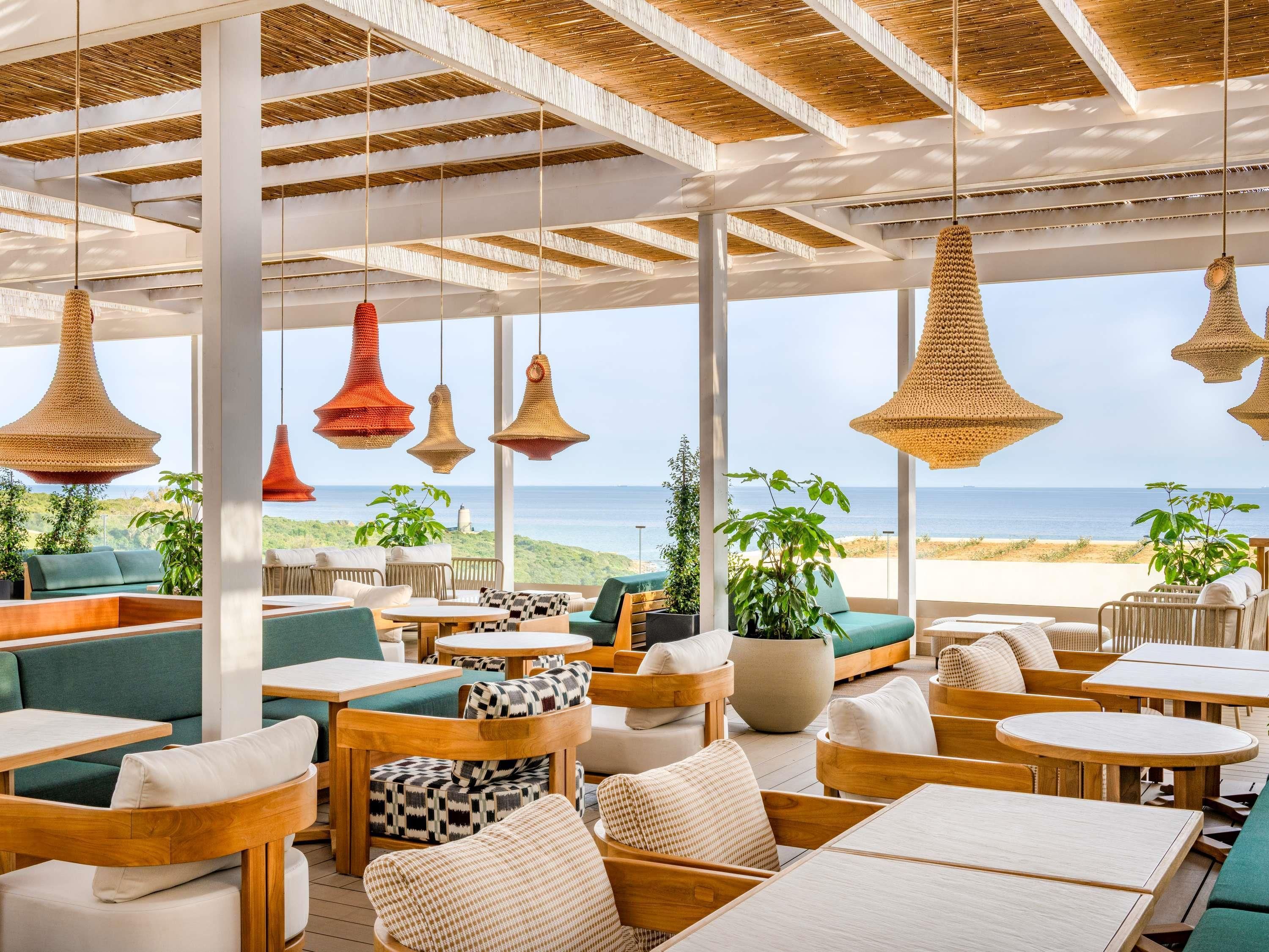 Un restaurante frente al mar con terraza, ventanas amplias y lámparas de mimbre colgantes.