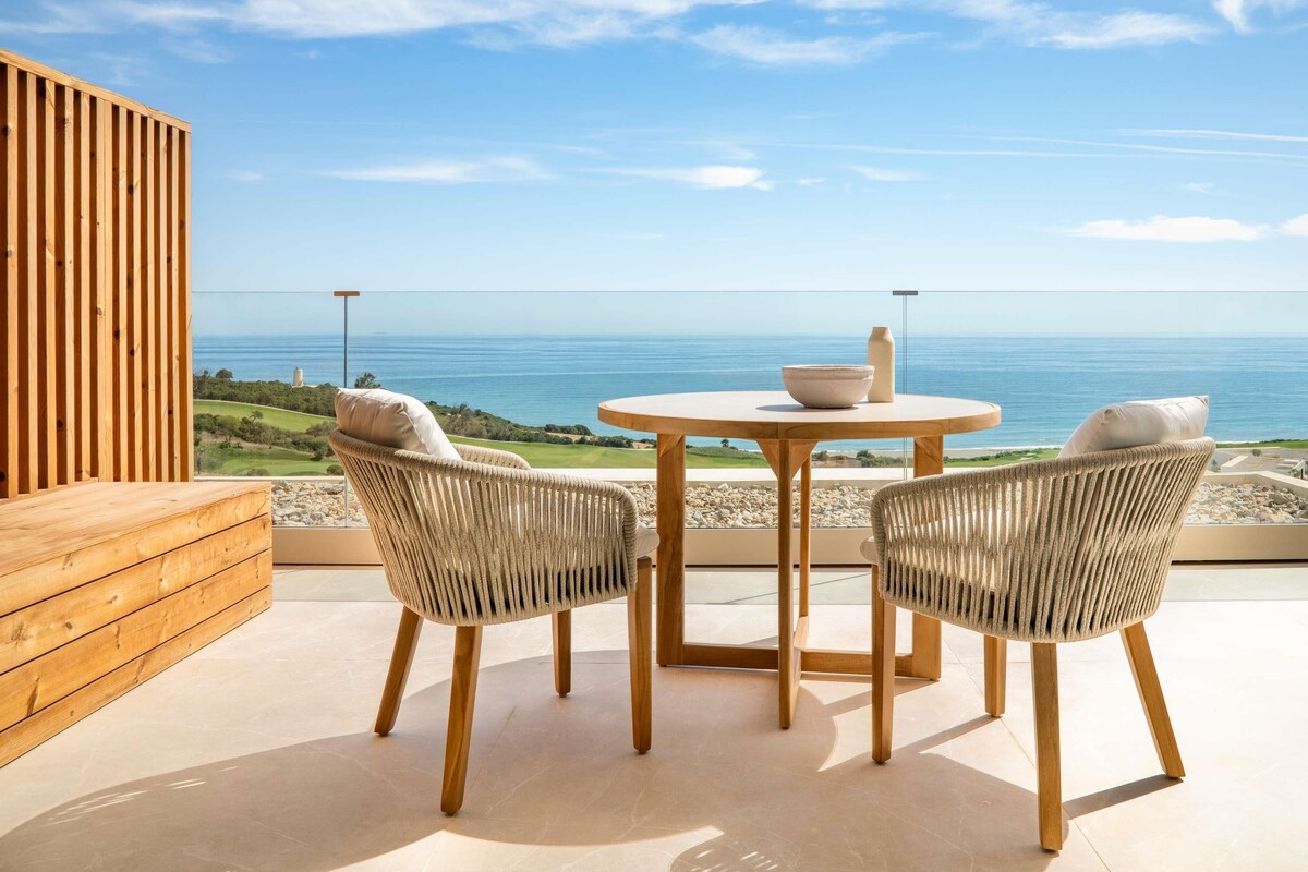 Une terrasse avec une table ronde et deux chaises en osier, donnant sur un paysage côtier.