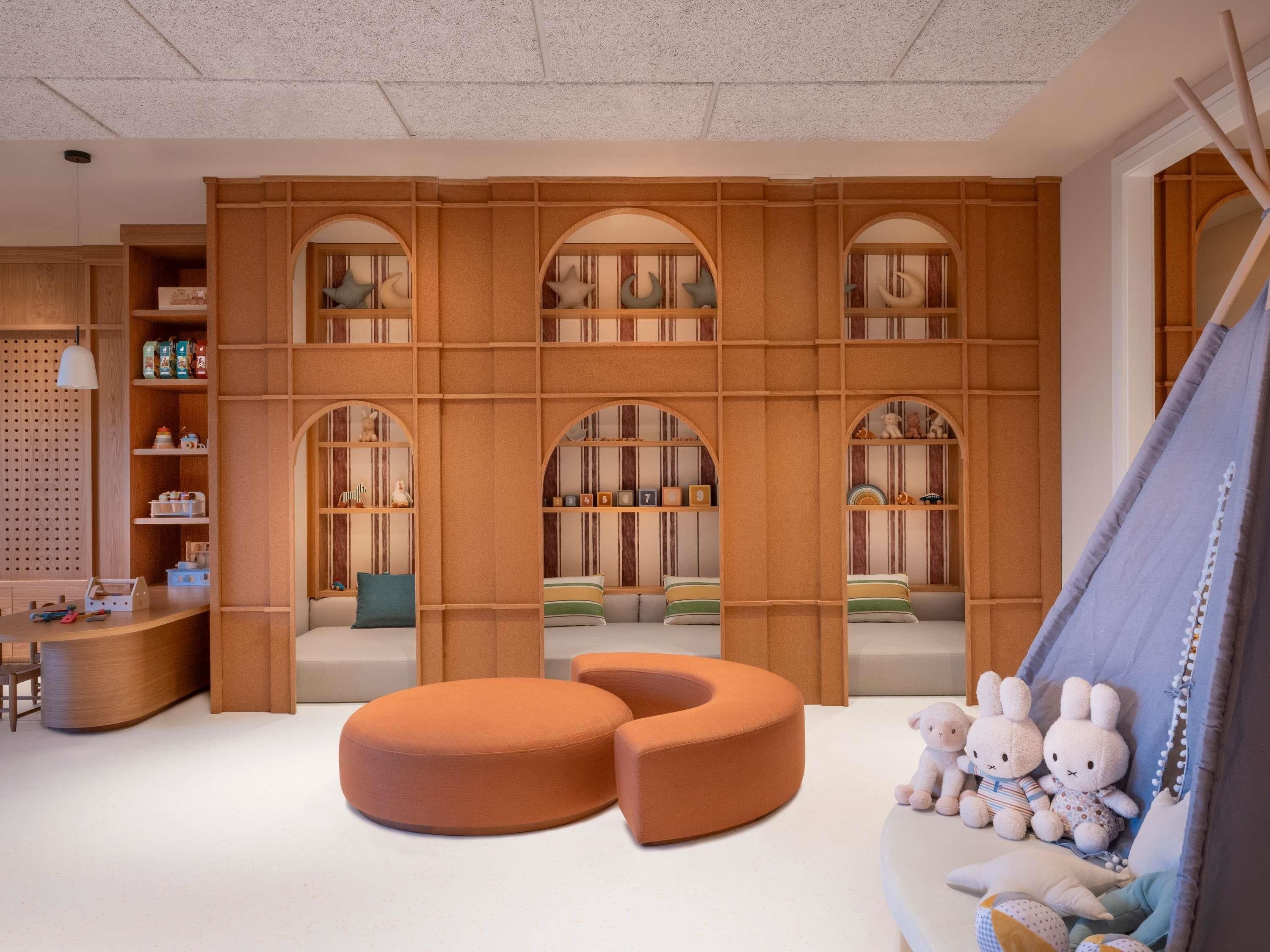 Una sala de juegos infantil con un otomano naranja grande, estantes con juguetes y una estructura parecida a una tienda llena de peluches.
