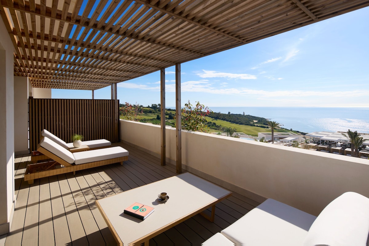 A balcony with a wooden pergola, a sofa, a table, and a view of the sea.