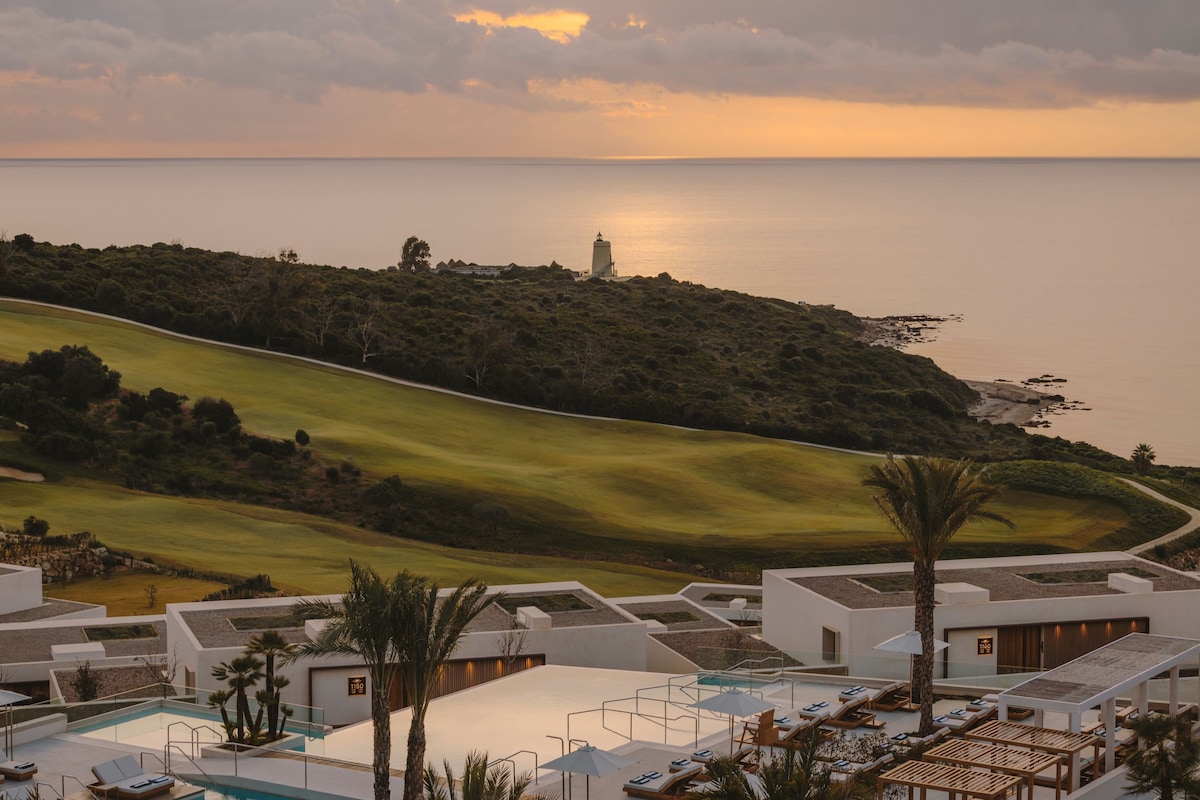 A resort with a swimming pool, sun loungers, and a view of the sea and a golf course.