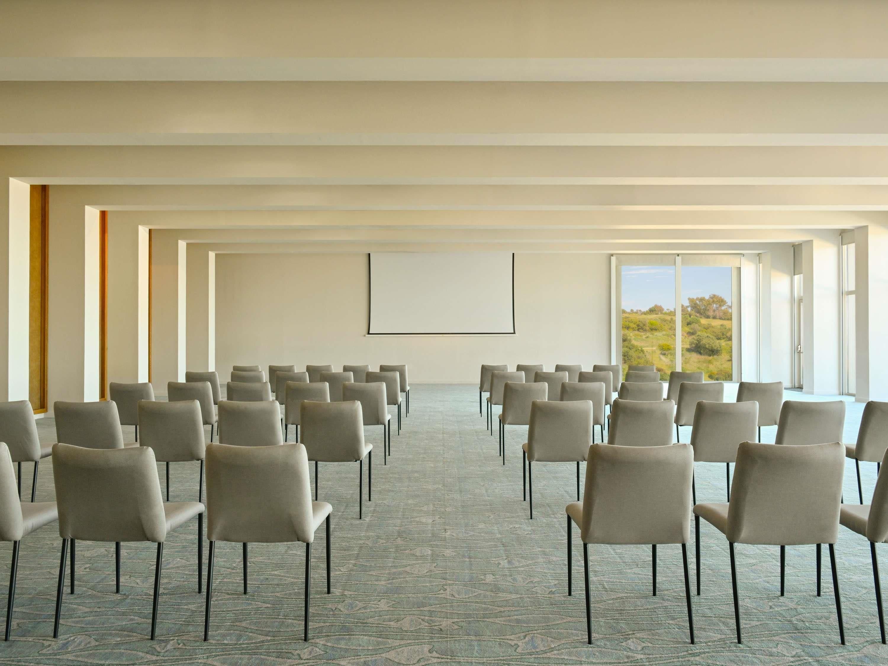 Un salón de conferencias con filas de sillas orientadas hacia una pantalla de proyección, ventanas amplias con vistas al exterior y suelo alfombrado.