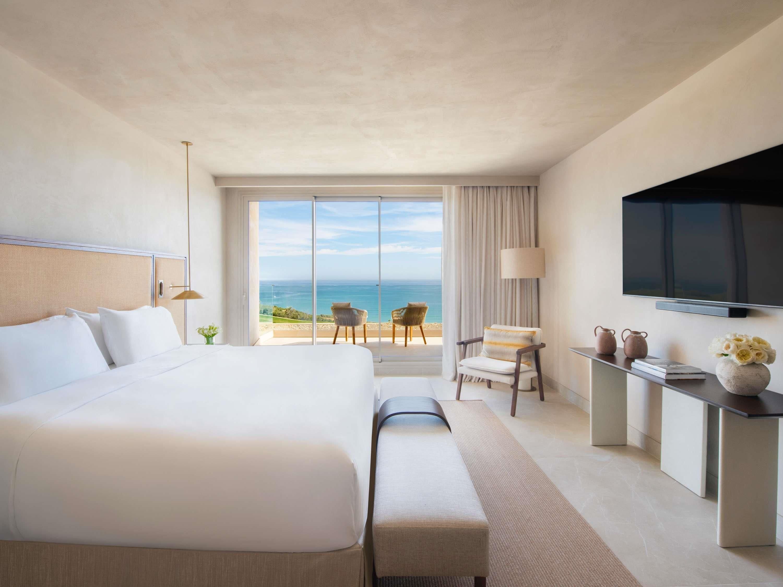 Habitación de hotel con una cama grande, televisión, escritorio con un florero y vista al océano.