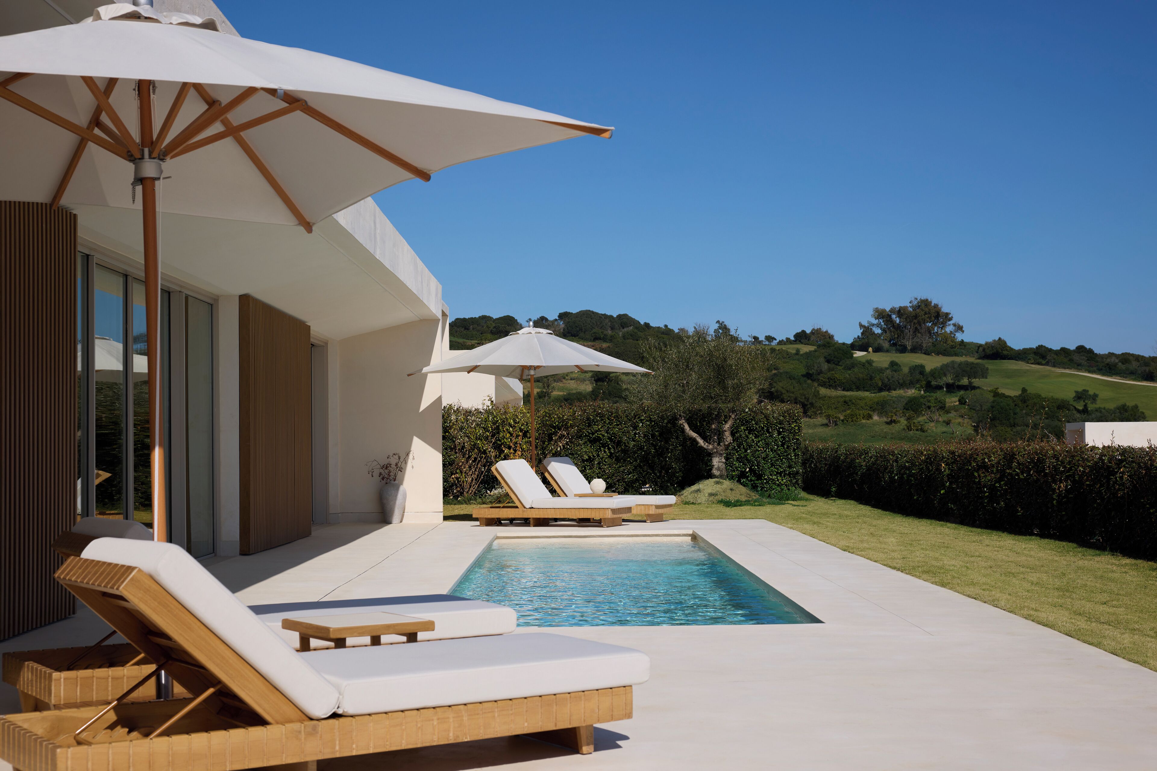Zona de piscina con sillones de descanso y sombrillas, rodeada de un césped cuidado y vistas a colinas onduladas.
