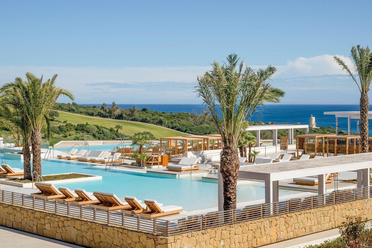 Un espace piscine agrémenté de chaises longues et de palmiers, offrant une vue sur un terrain de golf et l’océan.
