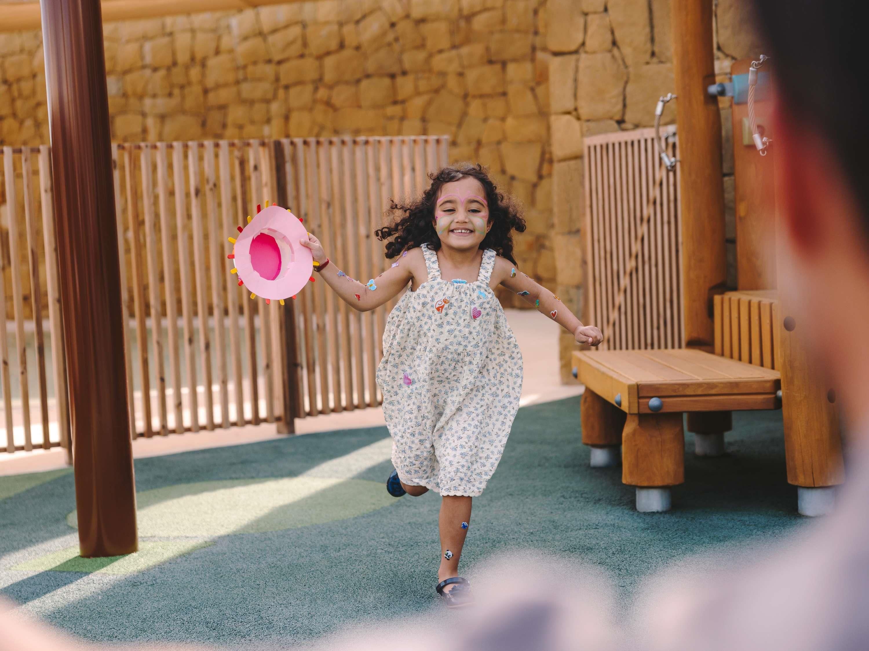 Un enfant court dans un parc, portant un chapeau rose, une robe à motifs floraux et un bracelet.