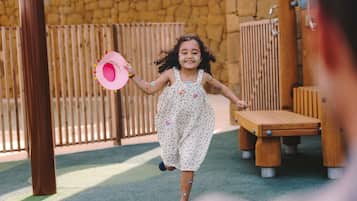 Un enfant court dans un parc, portant un chapeau rose, une robe à motifs floraux et un bracelet.