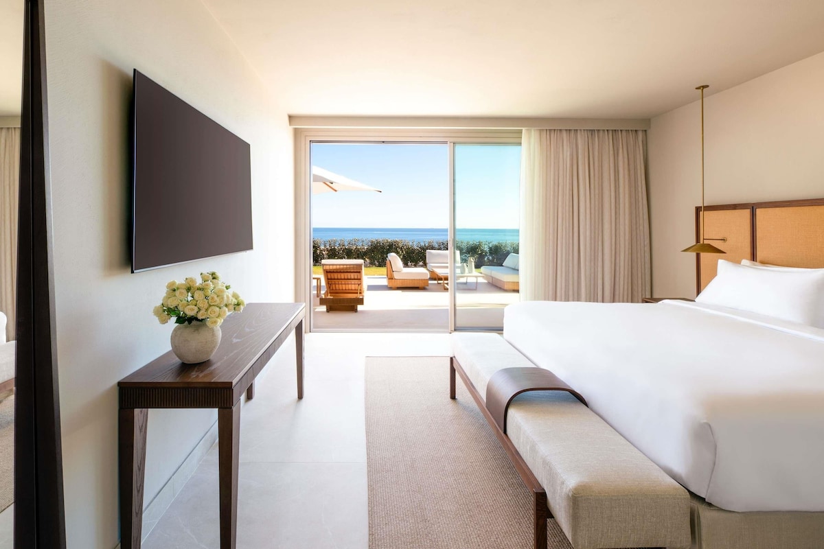 Ein Hotelzimmer mit Bett, Fernseher, einem kleinen Tisch mit Blumenvase, einer Bank und Meerblick durch eine Schiebetür.