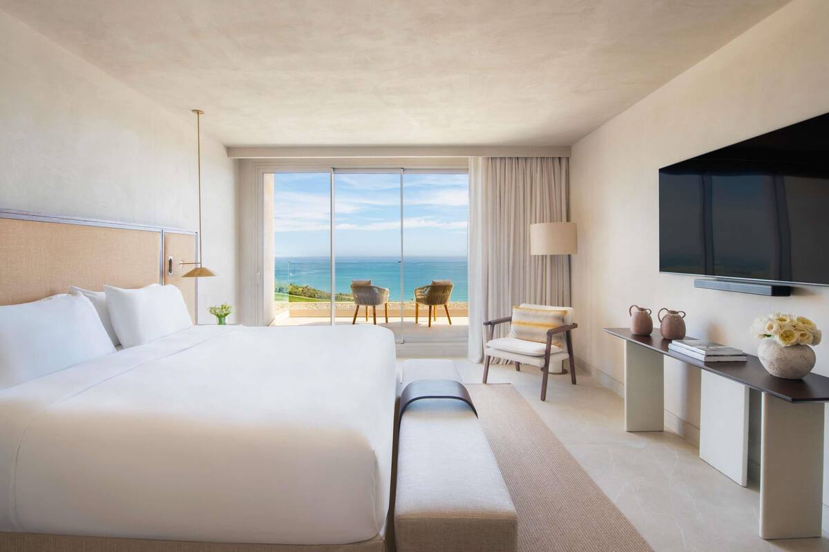 A hotel room with a large bed, a TV, a desk with a vase of flowers, and a view of the ocean.