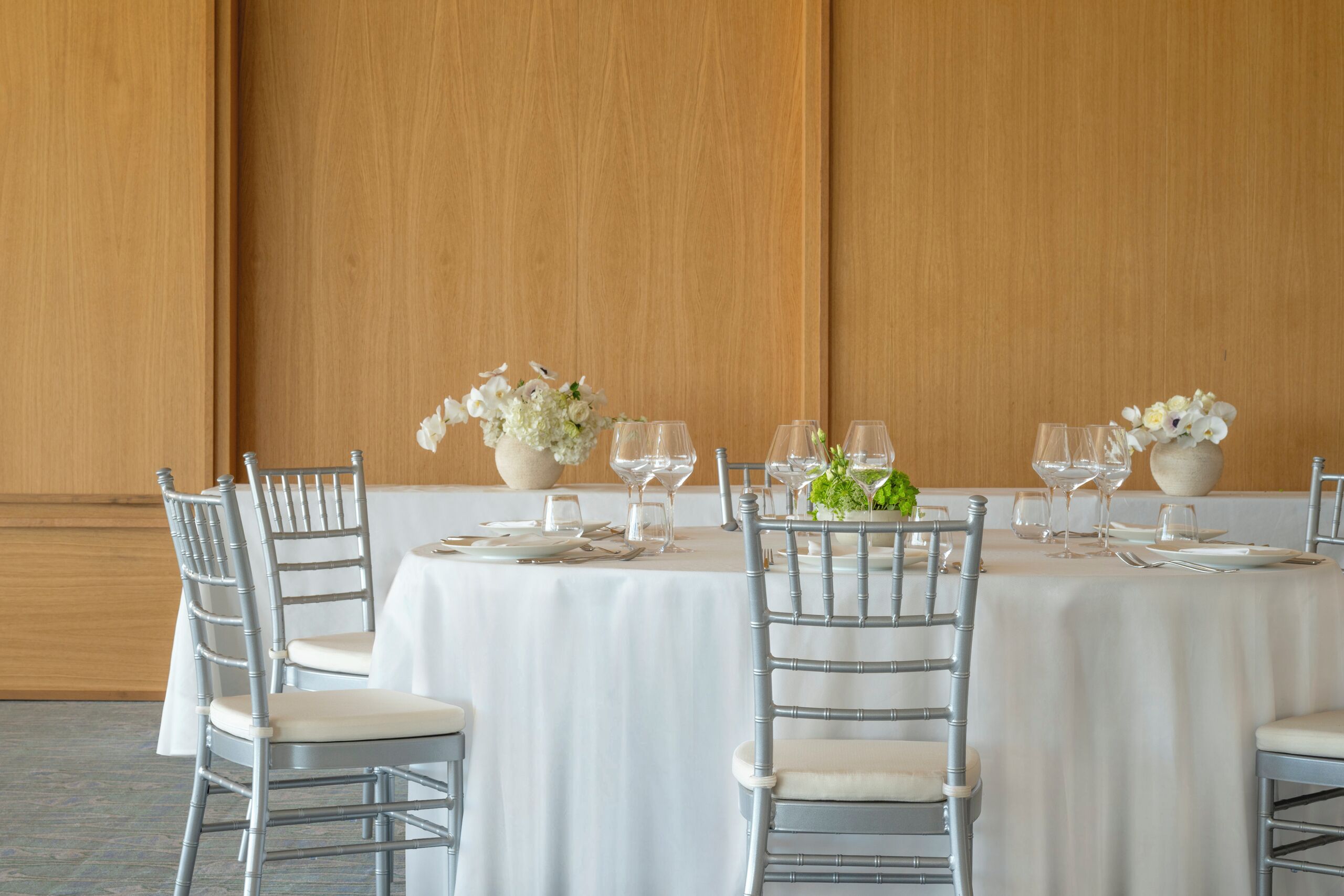 Un comedor con mesas redondas dispuestas para cuatro personas, cada una con mantel blanco, sillas de plata y copas.