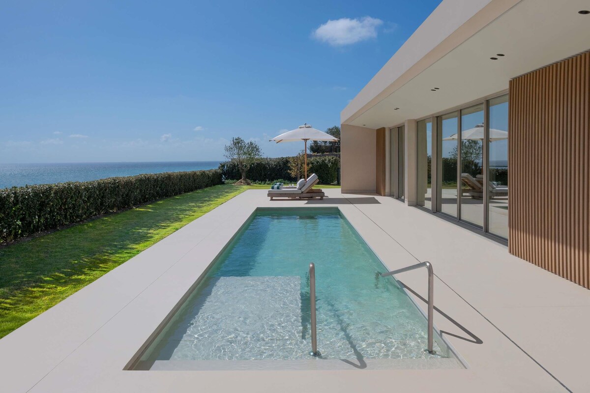 Une piscine extérieure moderne offrant une vue dégagée sur la mer, un espace détente avec des parasols et une pelouse bien entretenue.