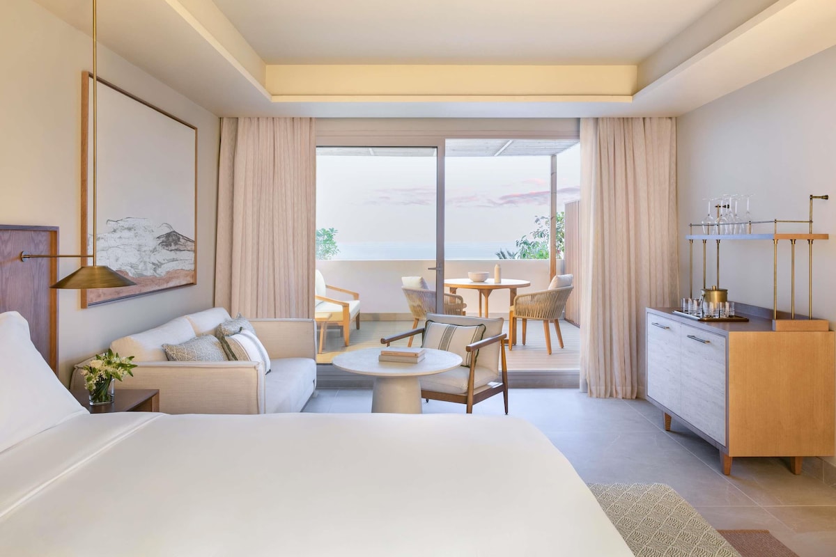 Une chambre d’hôtel moderne équipée d’un lit, d’un canapé, d’une table à manger, de chaises, d’un bureau et d’un balcon avec vue.