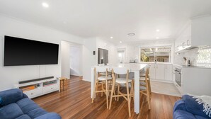 Un salon moderne doté d’un téléviseur à écran plat, d’un coin repas avec des chaises en bois et d’une cuisine avec des meubles blancs.