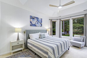 Een slaapkamer met een bed, nachtkastje, lamp, plafondventilator, raam met uitzicht op groen en een schilderij aan de muur.