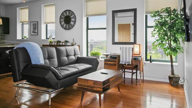 A modern living room with a black leather sofa, a wooden coffee table, and a large window with a view of the city. - Stylish Top Floor Condo in Downtown Indianapolis, Mile Square Neighborhood  (Indianapolis)