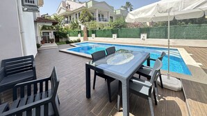 A swimming pool with a table and chairs, surrounded by a wooden deck and a green hedge.