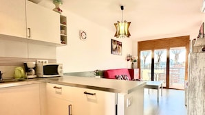 A modern kitchen with white cabinets, a microwave, and a coffee maker. There is a dining area with a red sofa and a table. A chandelier hangs from the ceiling, and a window offers a view of the outside. - V023 - Roses Platja (Roses)