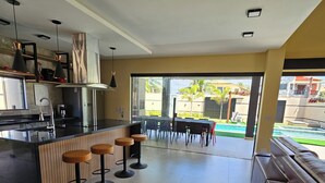 A modern kitchen with a central island, bar stools, and a dining area with a table and chairs. - Casa Luna (Camaçari)