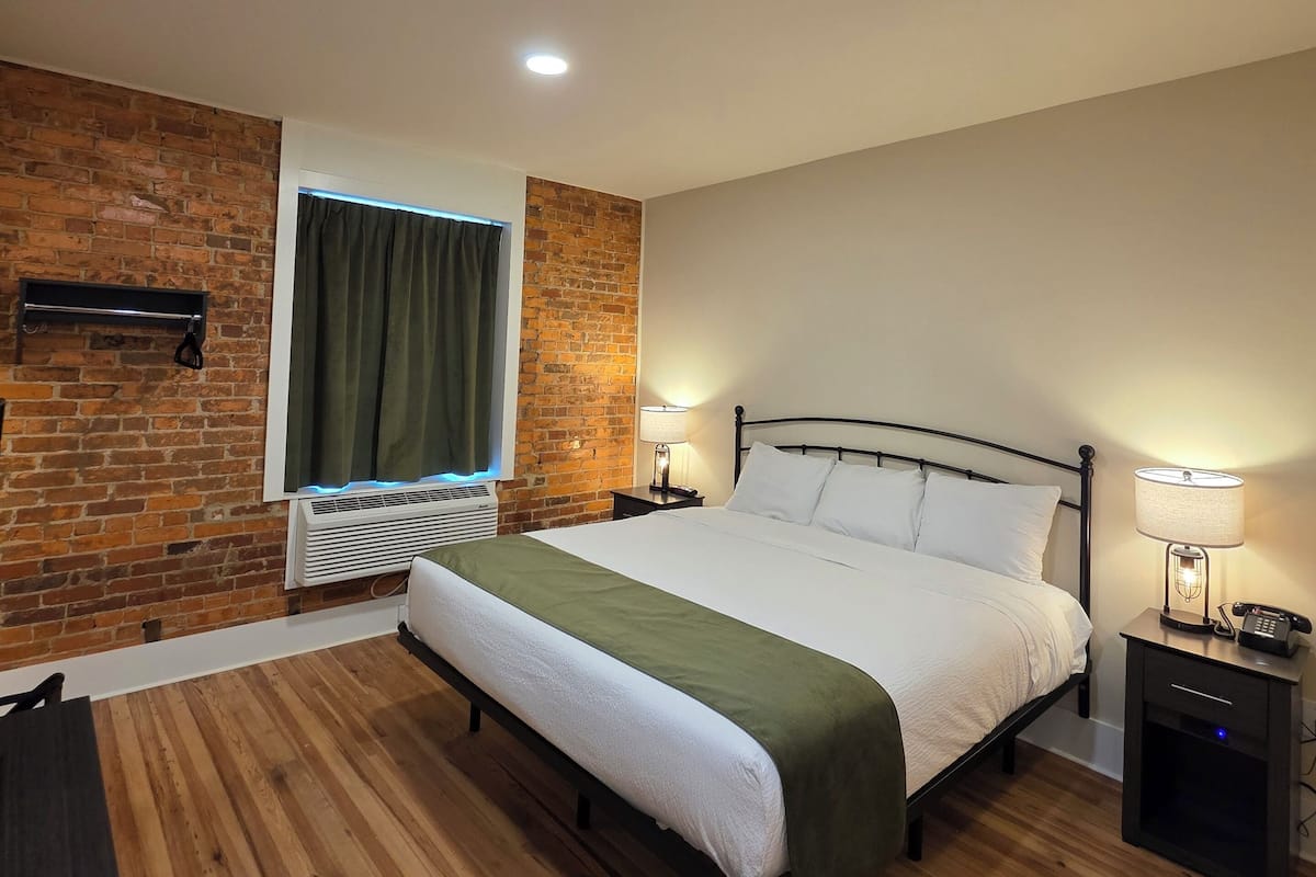 A modern hotel room with a brick accent wall, a large bed, a bedside table with a lamp, and a window with curtains.