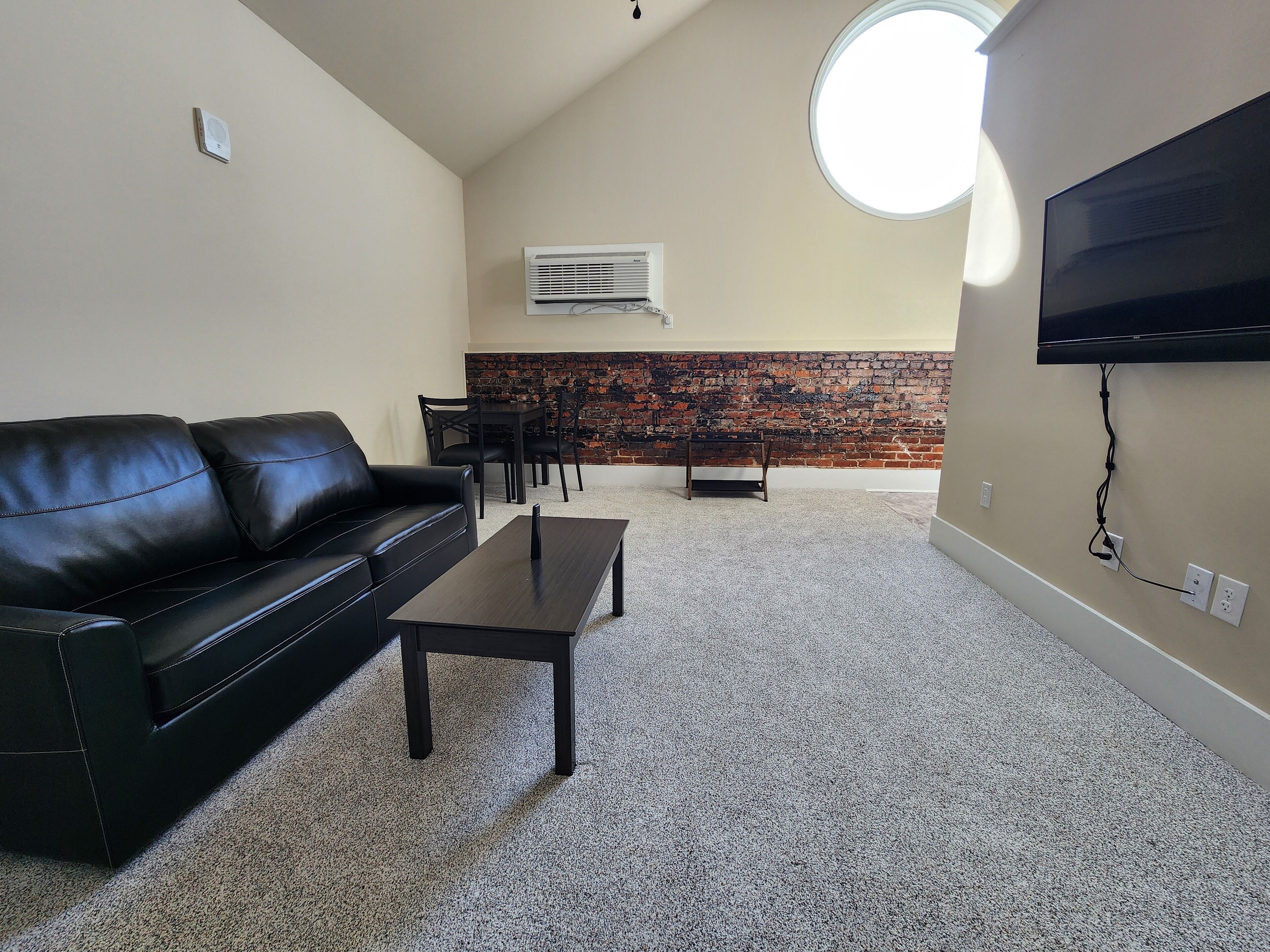 Una sala de estar con un sofá de cuero negro, una mesa de centro, un televisor montado en la pared, una pared de ladrillo decorativa y una zona de comedor con sillas y una mesa.