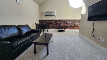 Una sala de estar con un sofá de cuero negro, una mesa de centro, un televisor montado en la pared, una pared de ladrillo decorativa y una zona de comedor con sillas y una mesa.