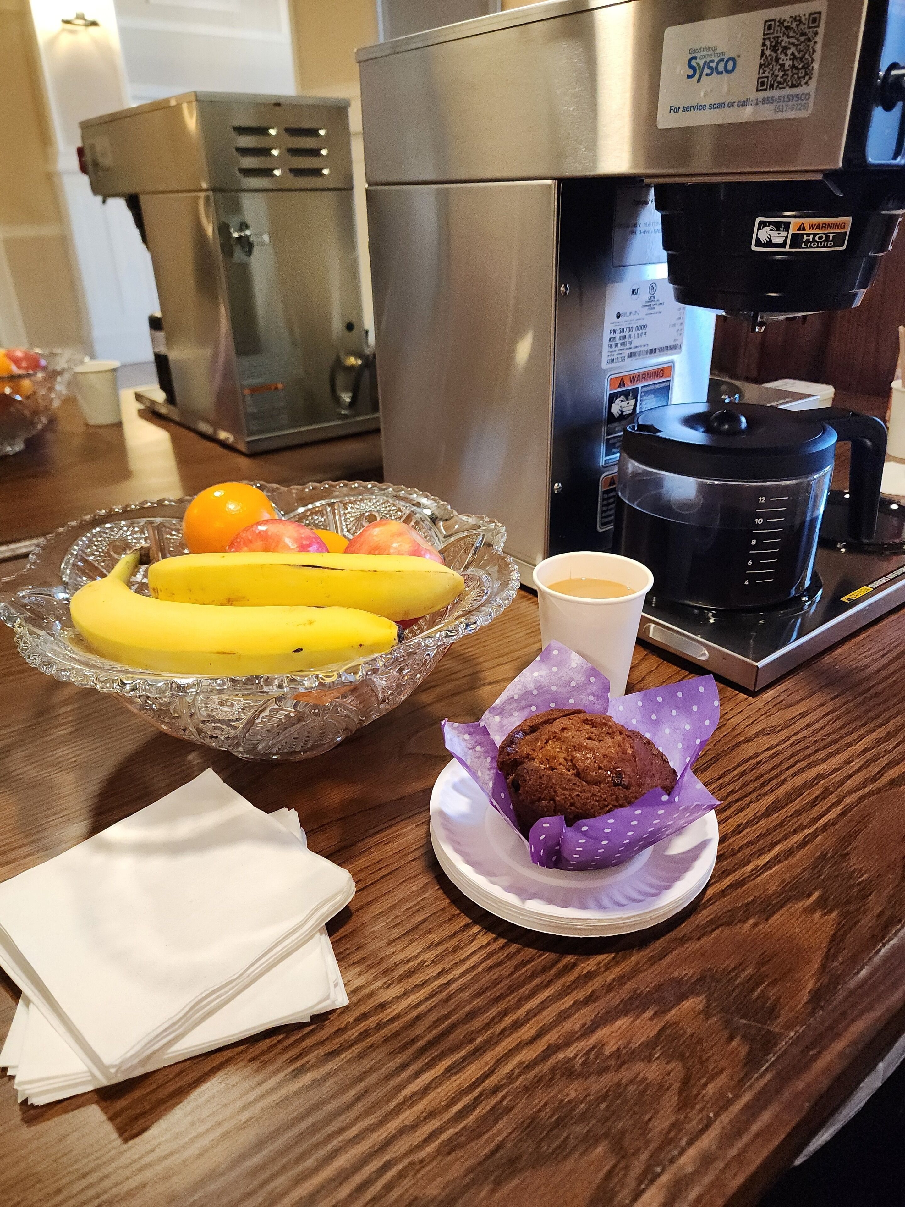 A kitchen counter with a coffee maker, a bowl of fruit, a muffin, and a napkin.