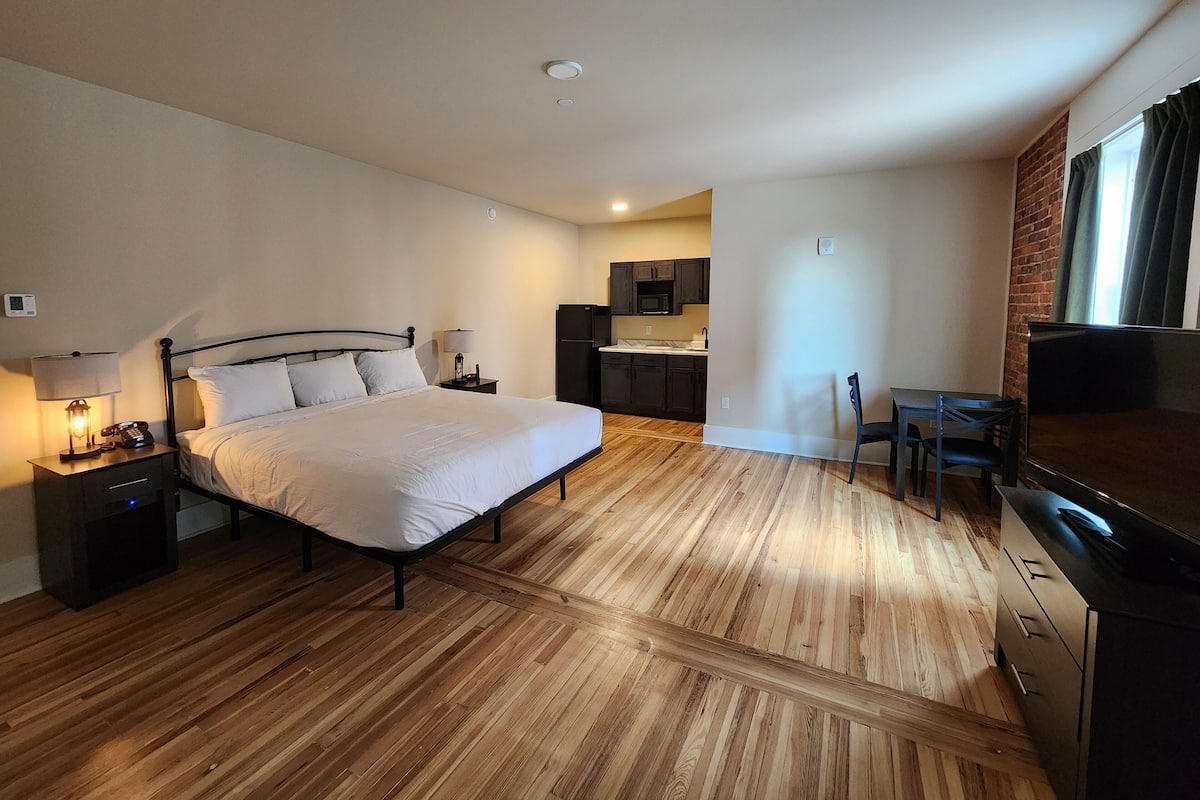 A hotel room with a bed, bedside tables, a desk, a chair, a TV, a kitchen area, and a brick wall.