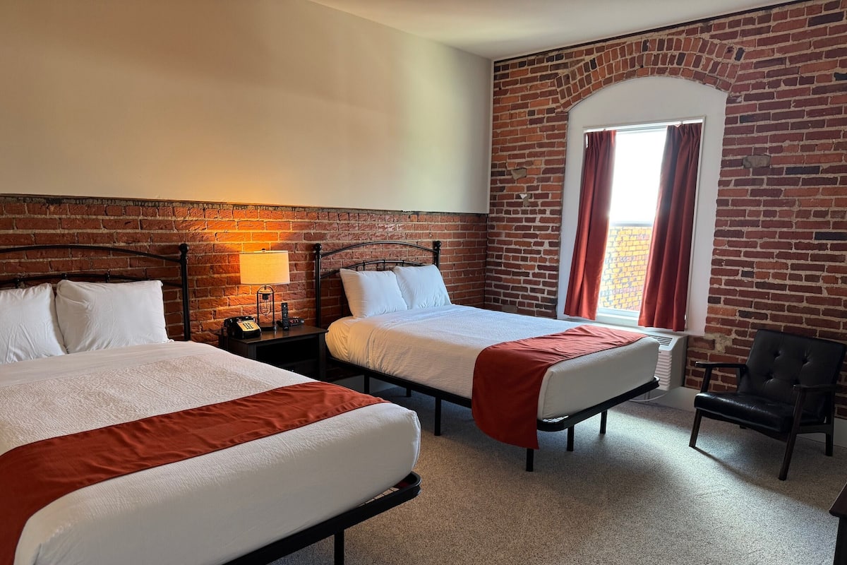A hotel room with two beds, a brick wall, a window with curtains, a chair, and a nightstand with a lamp and phone.