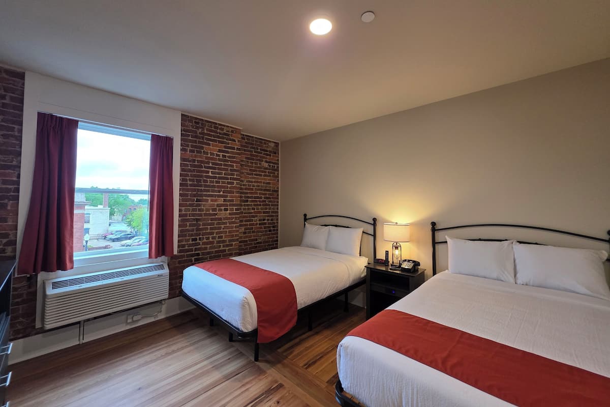 A hotel room with two beds, a brick wall, a window with curtains, a heater, and a view of the outside.