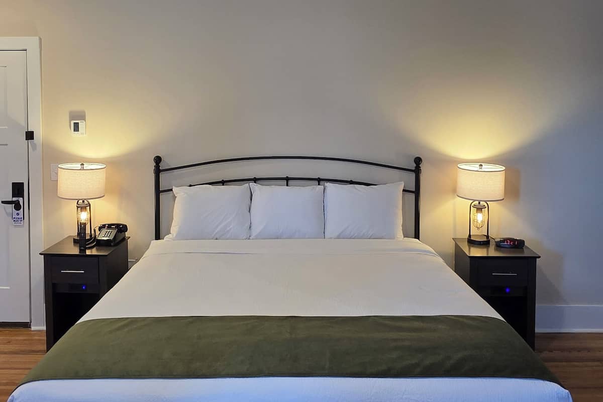 A neatly made bed with white linens and a green throw blanket, flanked by two bedside tables with lamps and clocks, in a room with wooden flooring and a white door.