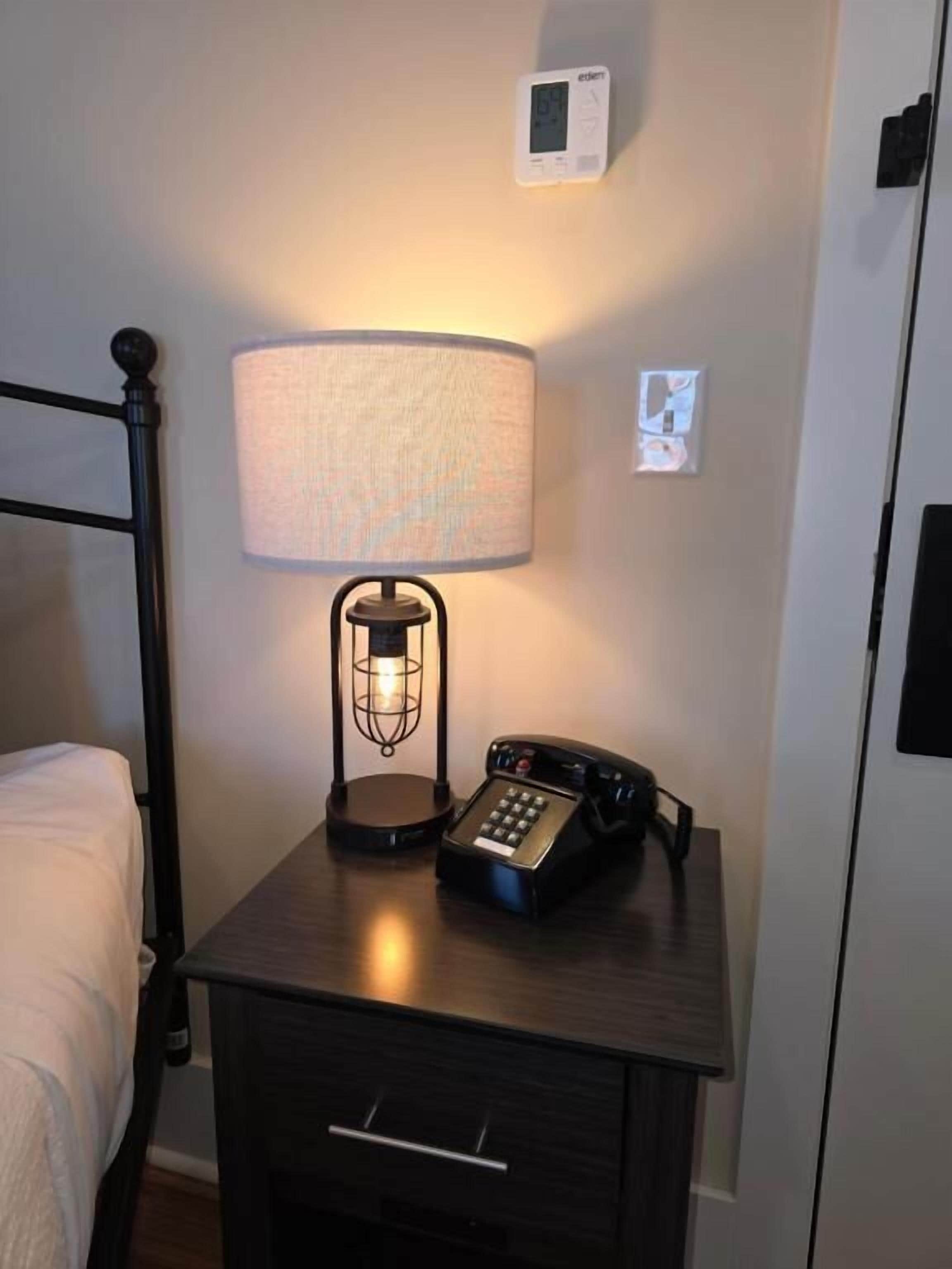 A hotel room with a bedside table featuring a lamp, a telephone, and a thermostat.
