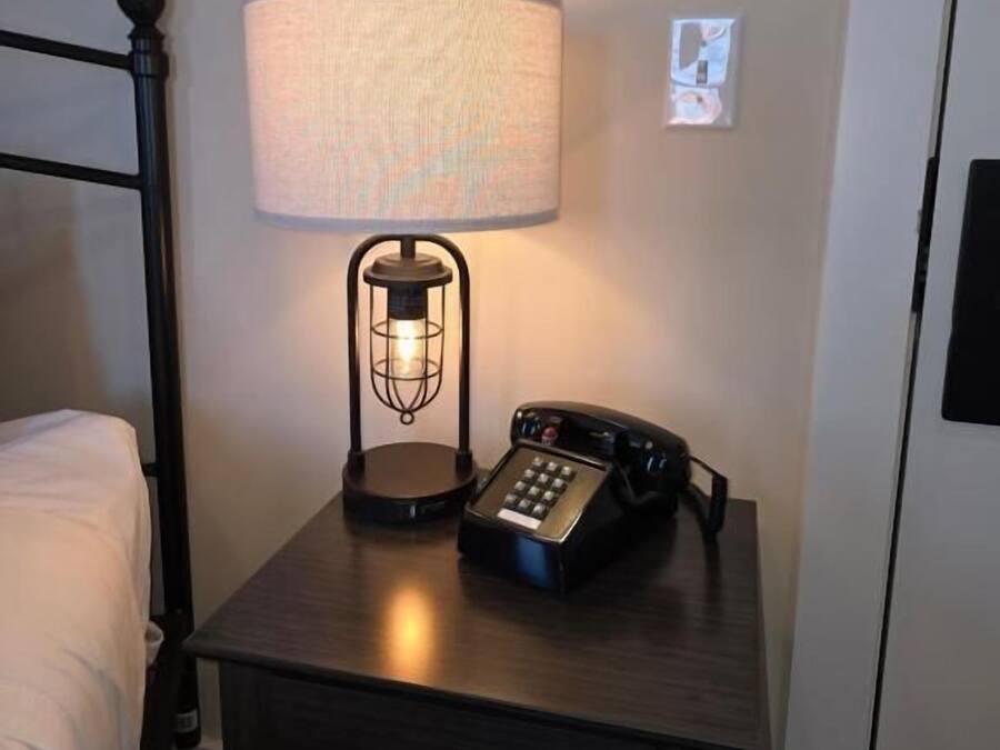 A hotel room with a bedside table featuring a lamp, a telephone, and a thermostat.