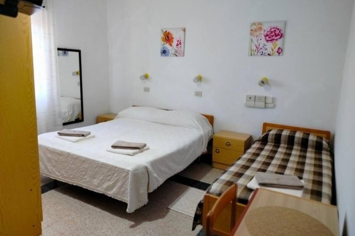 Quarto com uma cama de solteiro, uma pequena mesa de cabeceira, uma cadeira e um edredom xadrez. Há dois interruptores de luz e duas tomadas na parede.