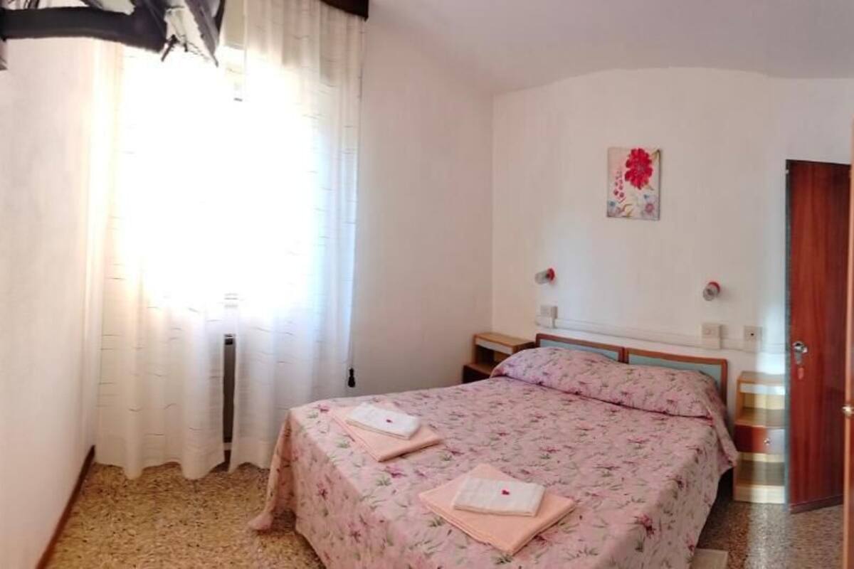 Quarto com cama, mesa de cabeceira, janela com cortinas, porta e luminária fixada na parede.