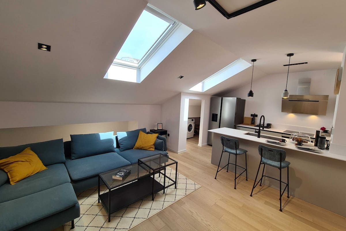 Ein modernes Loft-Apartment mit großem Dachfenster, einer Couch, einem Couchtisch, einer Küche mit Kühlschrank und einer Waschmaschine im Hintergrund.