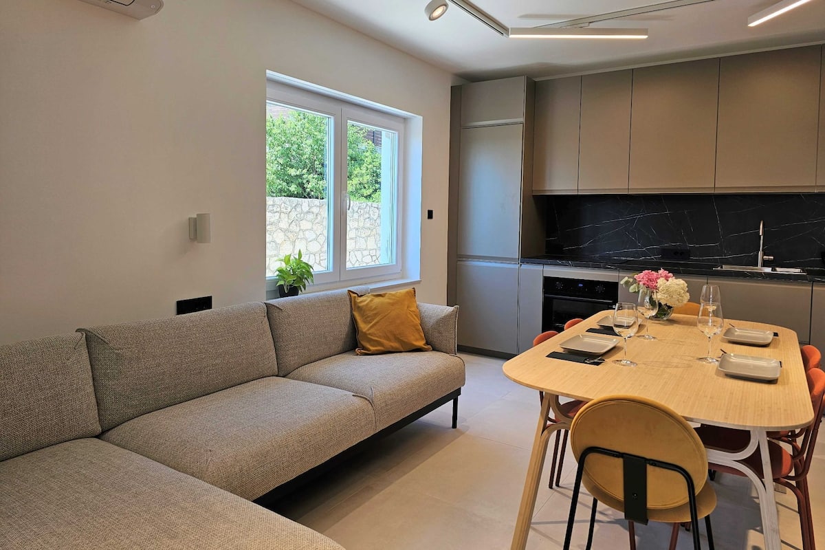 Ein modernes Wohnzimmer mit einer Couch, einem Esstisch mit Stühlen, einer Küchenzeile mit Schränken und einem Herd sowie einem Fenster mit einer Pflanze.