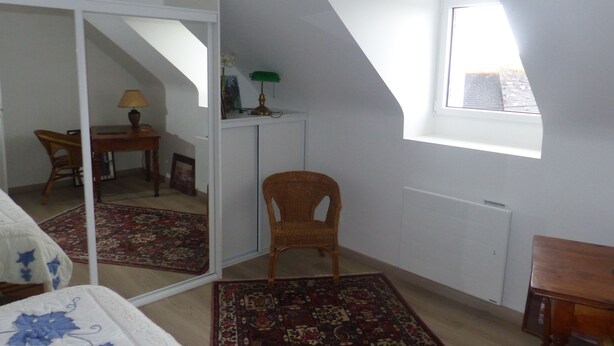 A room with a sloped ceiling, a small window, a mirror, a chair, a desk with a lamp, and a bed with a floral bedspread. - Fully renovated farmhouse in village near a typical fishing port (Hôpital-Camfrout)
