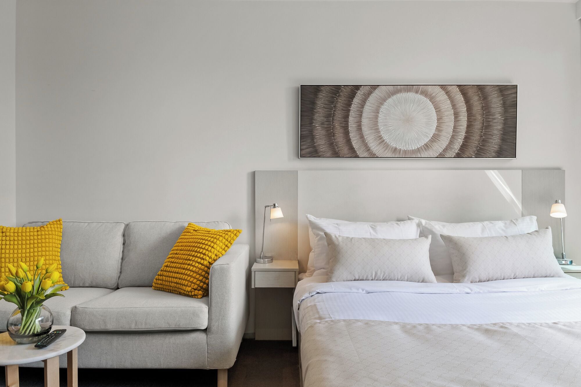 Une chambre d’hôtel avec un canapé gris, un lit avec une literie blanche, une œuvre d’art accrochée au mur et une table de chevet avec une lampe.