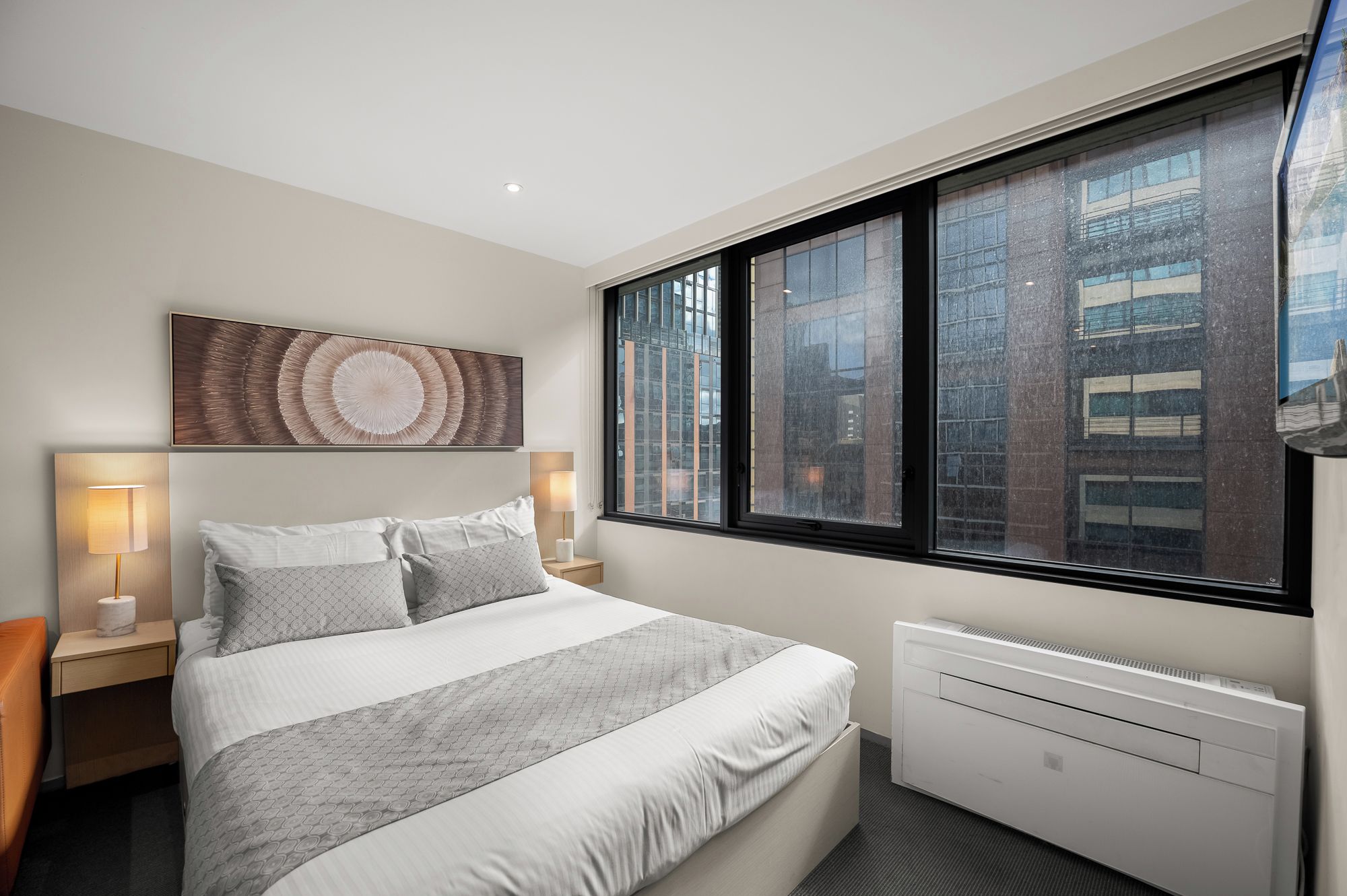 A modern hotel room with a large bed, two bedside lamps, a radiator, and a view of a cityscape through large windows.