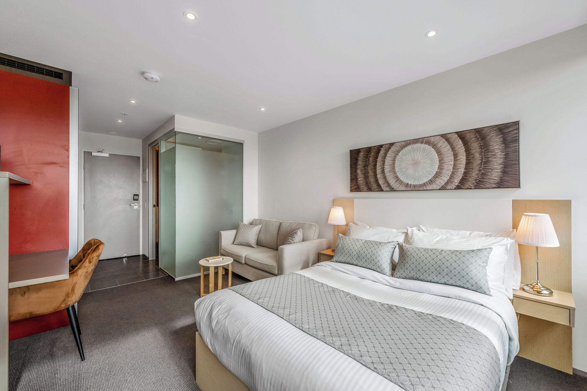 Une chambre d’hôtel moderne avec un grand lit, deux tables de chevet avec des lampes, un canapé et un mur d’accent rouge.