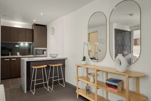 A modern kitchen with dark cabinets, a microwave, and a sink. There are two wooden stools and a white console table with books and a vase. - City Stays (Melbourne)